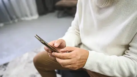 Getty Images A close up on a man's hands using a mobile phone. He is wearing a cream jumper and brown trousers and sitting in a room with a grey rug and carpet.