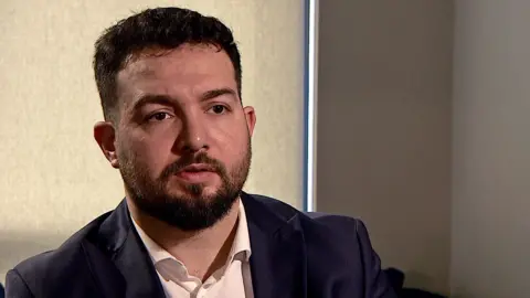 BBC A man in a white shirt and black suit jacket sitting in a darkened room with the blind drawn. He has dark short hair and a dark beard.